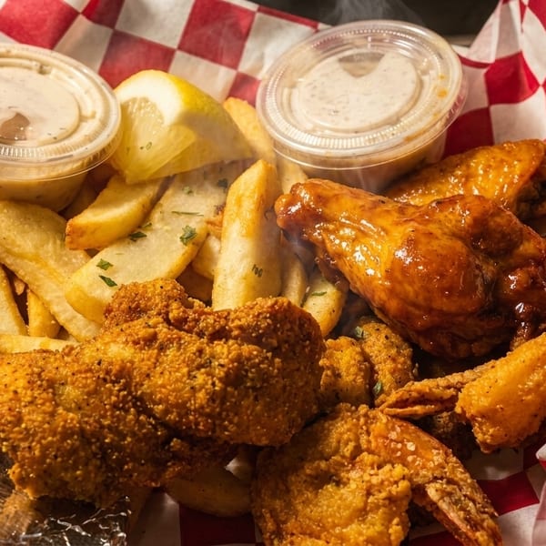 Steaming hot fried chicken, shrimp, and fries with dipping sauces and lemon in a red checkered basket.